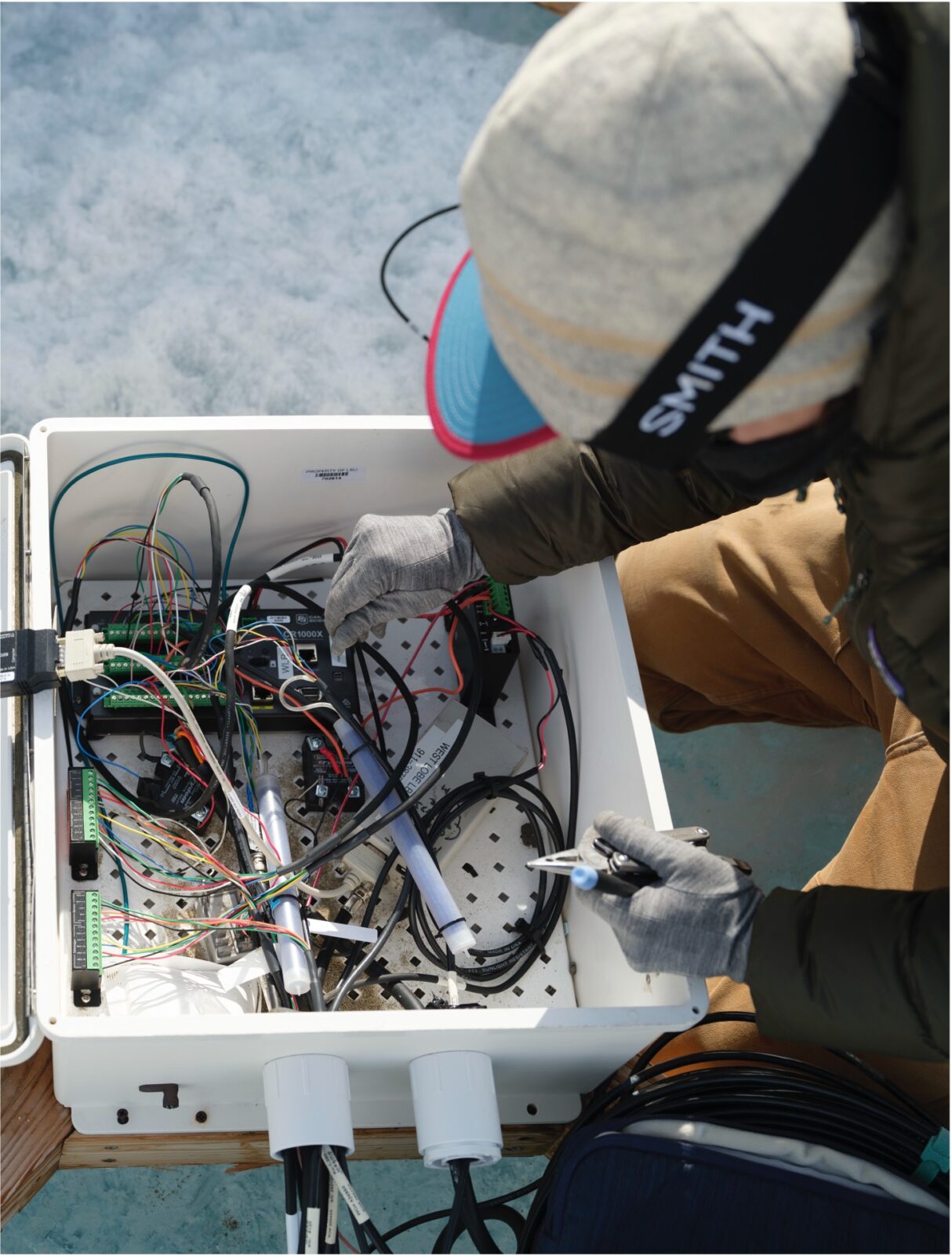 A photo looking over someone's shoulder as they investigate wiring inside of a small white box on top of frozen ground.
