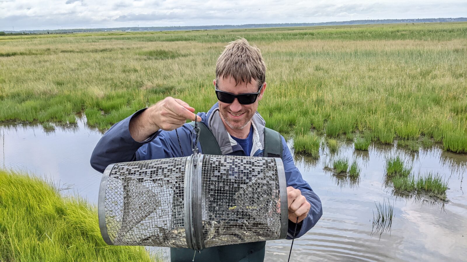 A photo of a person standing near a stream holding a metal trap dripping with water.