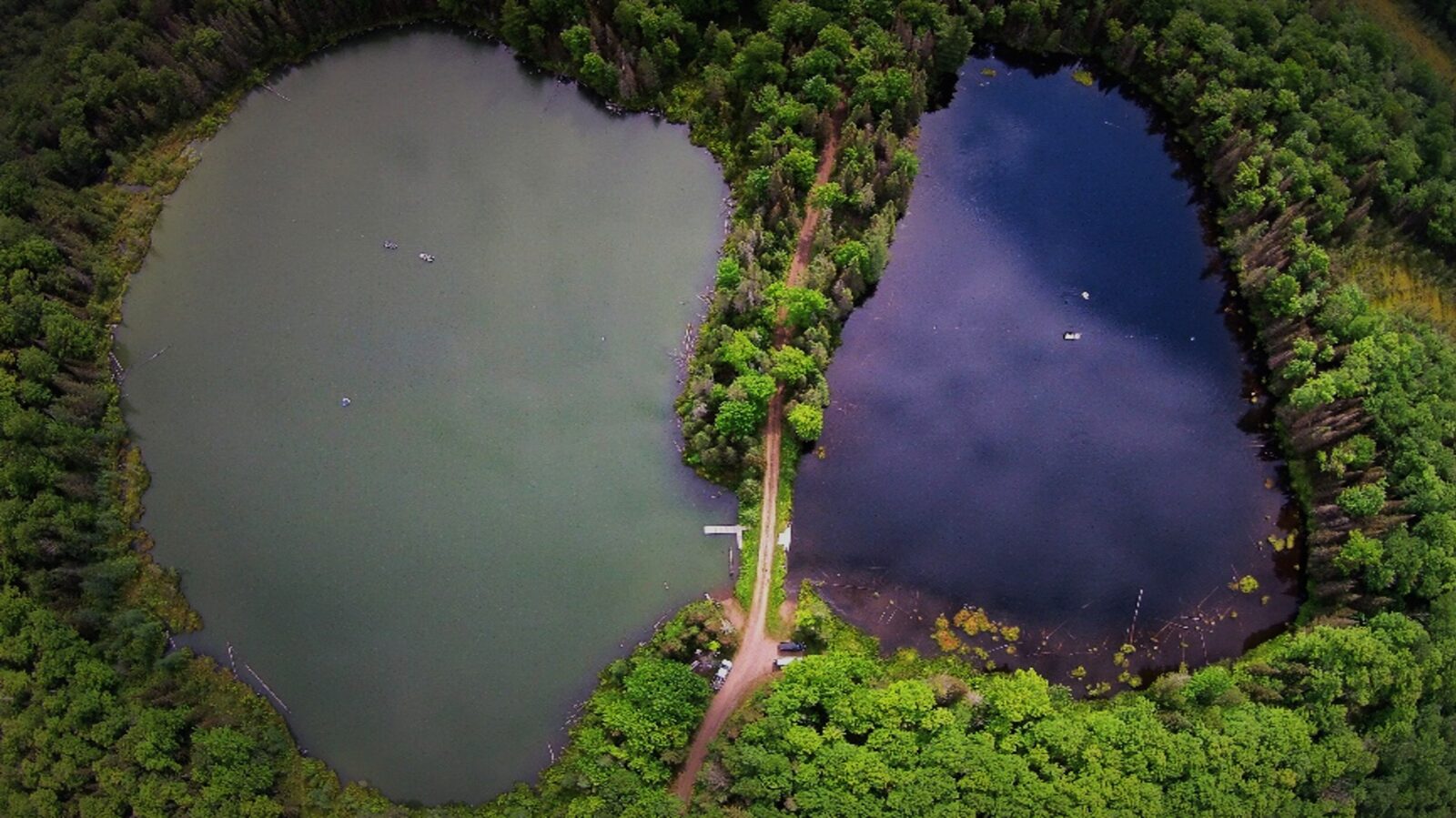 An aerial photo of two lakes divided by a narrow road and surrounded by forest. The lake on the left appears green, while the lake on the right looks more blue.