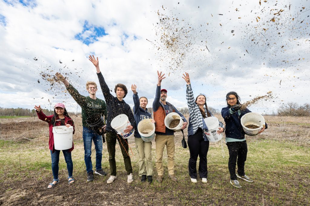 A line of people in a field have one arm thrown up into the sky, releasing the handful of seeds they just gathered from the large white buckets held in their other arm.