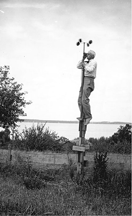 A historic photo of a man standing on top of a post while reading data from a weather station. A lake lies in the background, and the post the man is standing on rises in an overgrown grassy field.