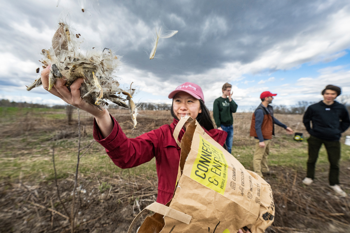 A young adult in a red jacket and pink baseball cap smiles at the fist full of fluffy seeds she holds up to be caught in and carried by the wind. They stand in a field of short green-brown grass that is patched with burn marks from a previous prescribed burn.
