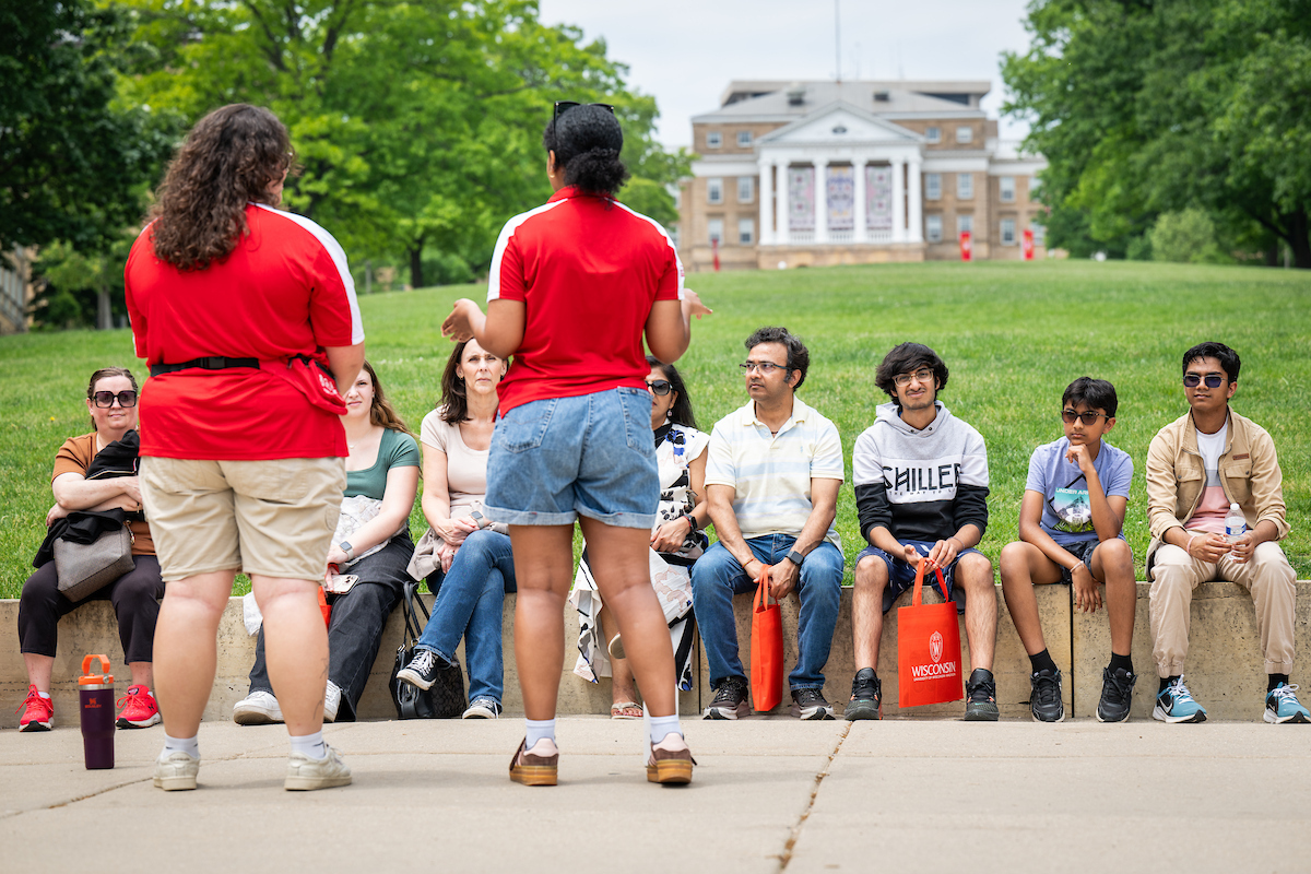 A group of prospective students and their families sit at the bottom of Bascom Hill looking at two tour guides in bright red collared shirts as they give them a tour of campus.