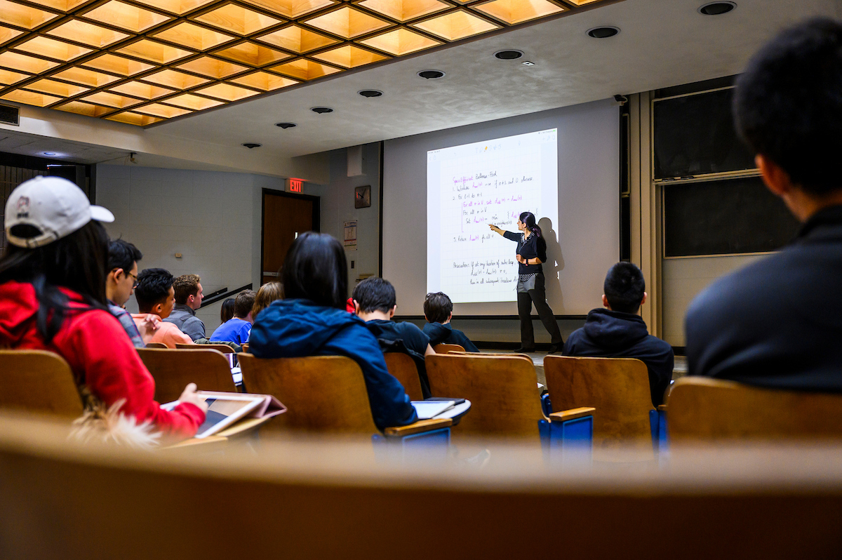 A professor points to a particular line of their presentation during a lecture in a large lecture hall while students watch and take notes from the audience.
