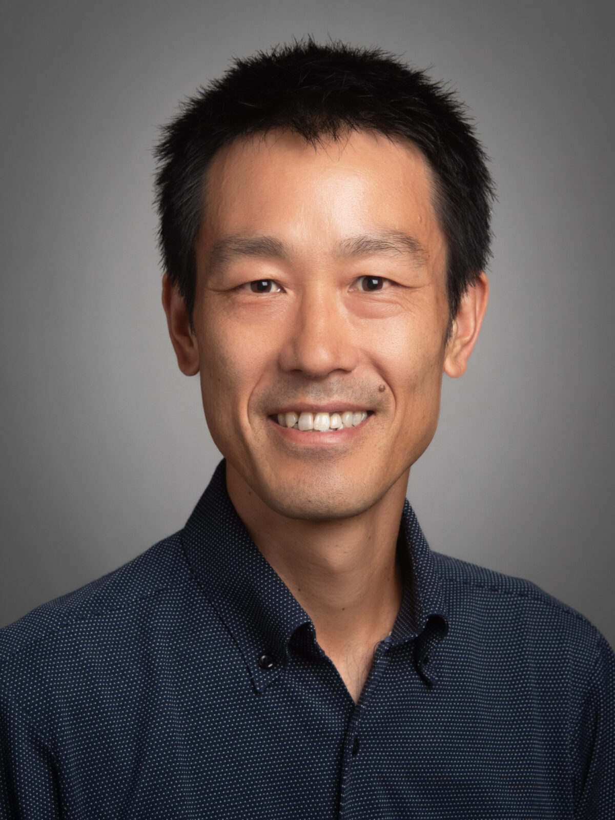 A studio portrait of a smiling person in front of a grey background.