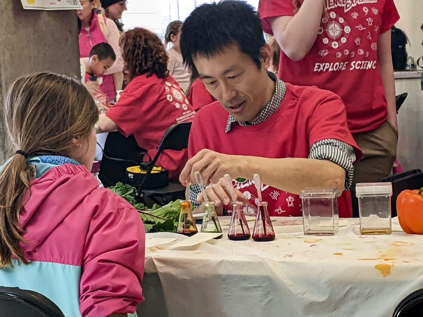A professor in a red tshirt branded with "Science Expeditions" logo, picks up a pipette to show the interested child across the table an experiment with plan pigments.