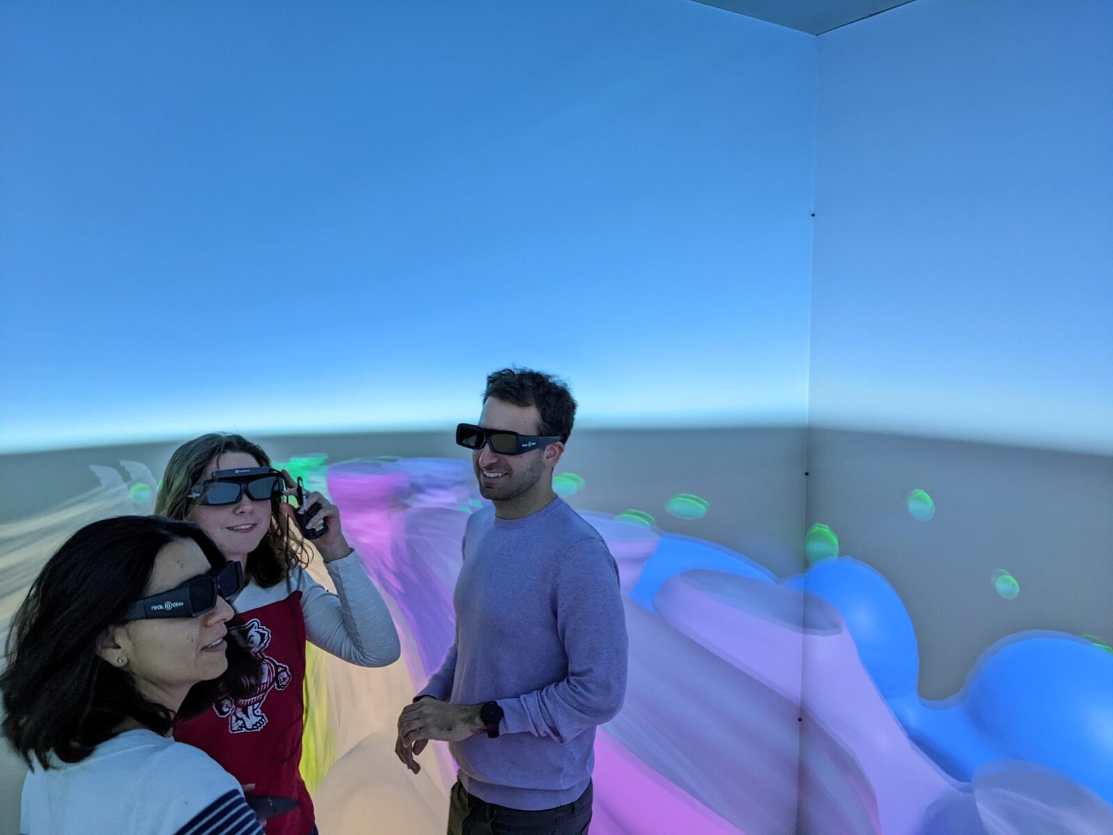A photo of three people wearing large glasses and looking at a colorful landscape projected onto the walls around them.