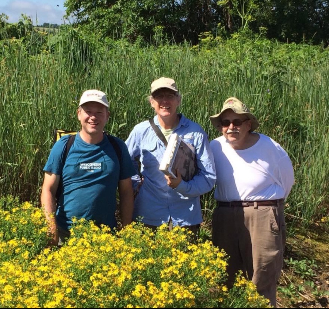 Three people stand in a field and smile for a photo surrounded by tall grasses behind them and a bush of bright yellow flowers in front of them.