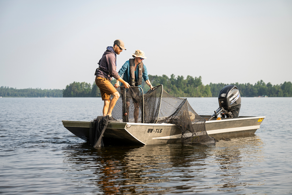 Two people wearing hats and slim life vests stand on a small aluminum boat floating the a lake. They both maneuver and guide a long stretch of net with cylindrical framing into the water alongside the boat.