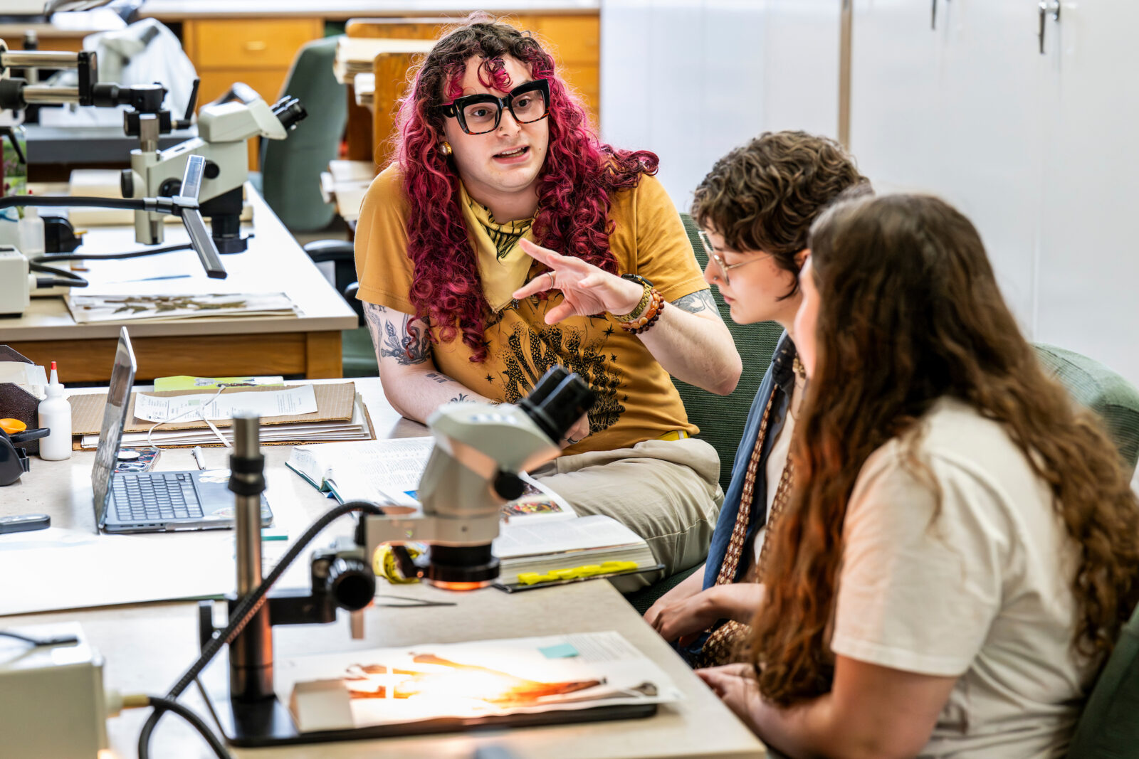 A group of three sit in a row down the length of a table strewn with books and other materials. The two closest to the camera are turned away and facing the third person who has bright pink curly hair and uses their hands to emphasize a point as they talk with the other two.