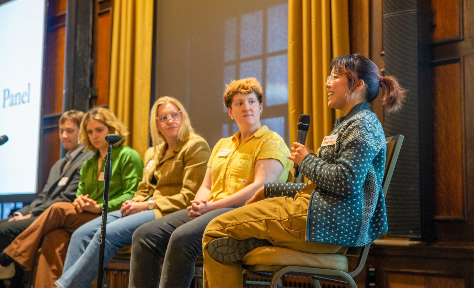 A panel of people sits in a row looking down the line to the panelist at the far end of the line holding a microphone and speaking out to the audience.
