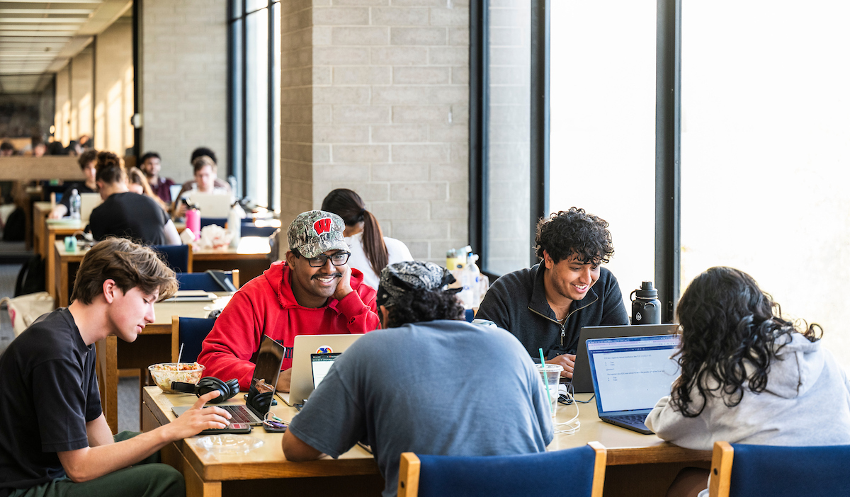 A group of students sit around a library table near large windows, studying on laptops and with notes.