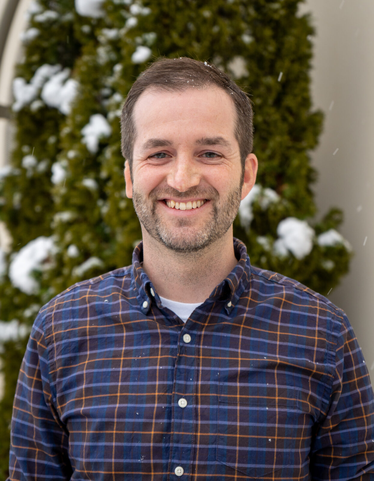 A portrait of a person smiling with an evergreen tree with snow on it in the background and small snowflakes falling around them.