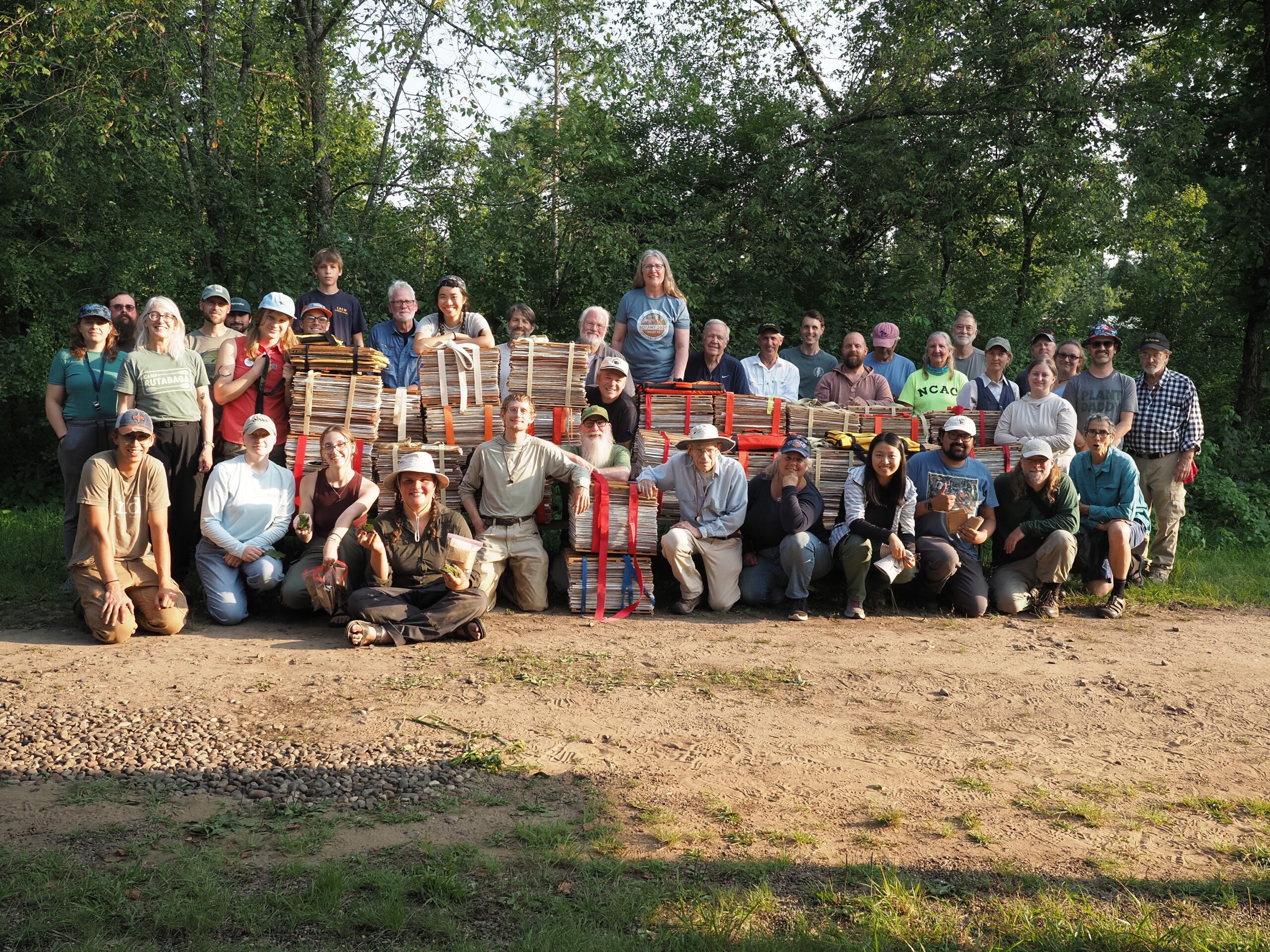 A large group of people pose for a photo together outside around large stacks of pressed plants.
